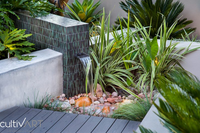 water feature with mosaic tiles and stainless steel water spout designed as part of composite decked area with washed river stones and dado finish wall
